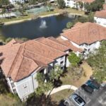 Aerial view of an apartment complex with red-tiled roofs, a small pond with a fountain, nearby tennis courts, and parked cars along a curved driveway surrounded by trees.