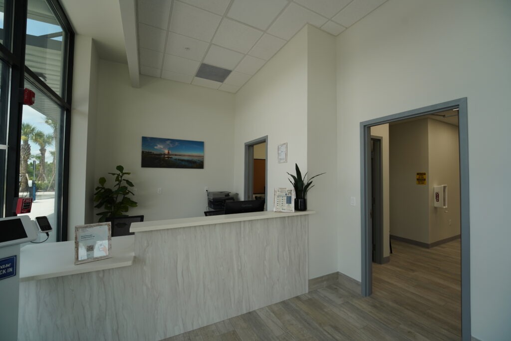 DSC00087 A modern, clean reception area with a white counter, potted plants, and a computer. Artwork hangs on the wall behind the desk. The space features large windows, light wood flooring, and doorways leading to other rooms.
