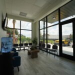 A waiting room with chairs along the walls, a water cooler, a coffee table with plants, and large windows showing parked cars and trees outside on a sunny day.
