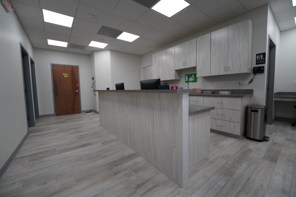 DSC00106 A modern, clean medical office reception area with a wood-textured counter, gray flooring, white cabinets, a computer, and a closed wooden door in the background under bright ceiling lights.