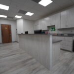 A modern, clean medical office reception area with a wood-textured counter, gray flooring, white cabinets, a computer, and a closed wooden door in the background under bright ceiling lights.