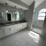 Modern bathroom with white cabinets, a large oval mirror, marble countertops, pendant lights, and a walk-in glass shower. Sunlight enters through a tall arched window, casting reflections on the shiny tile floor.