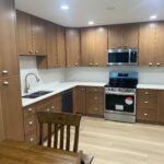 Modern kitchen with wood cabinets, stainless steel appliances, white countertops, a double sink, and a small dining table with a wooden chair in the foreground. Bright overhead lighting illuminates the space.