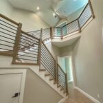 Modern staircase with wooden handrails and black metal balusters, leading to an upper floor with a landing. Walls are painted light beige, and the space is well-lit by ceiling lights. Doorways visible on both floors.
