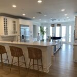 Modern kitchen with white cabinets, a large island, wicker barstools, stainless steel appliances, and wood flooring. Sunlight streams in through glass doors at the back, and a green plant sits near the windows.
