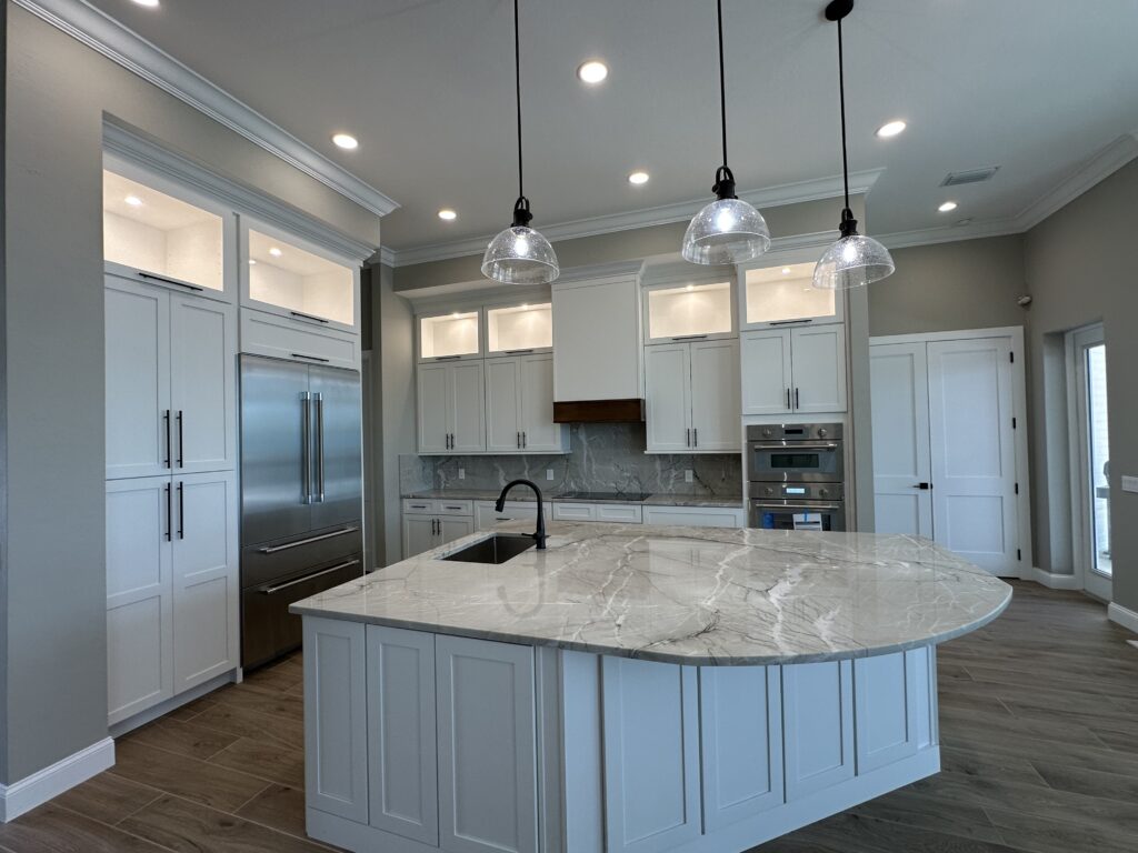Modern kitchen with white cabinets, stainless steel appliances, a marble island with a built-in sink, pendant lights overhead, and wood-look tile flooring. The space is bright and open with recessed lighting.