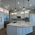 Modern kitchen with white cabinets, stainless steel appliances, a marble island with a built-in sink, pendant lights overhead, and wood-look tile flooring. The space is bright and open with recessed lighting.