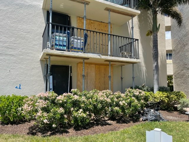 Loggerhead Concrete A two-story building with balconies under renovation, supported by scaffolding and metal poles. As a concrete restoration contractor in Florida, the site features boarded-up lower windows and doors, with flowering bushes and green grass in front.