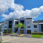 A modern, light gray apartment building with dark roof tiles, balconies, and palm trees in front, under a bright blue sky with scattered clouds. A driveway and parking area are visible in the foreground.