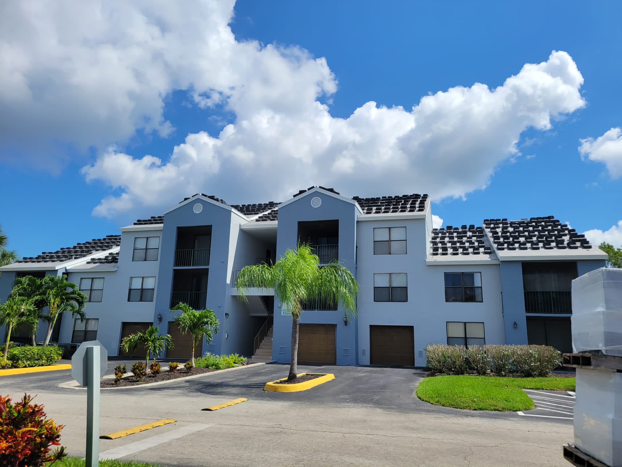 A modern, light gray apartment building with dark roof tiles, balconies, and palm trees in front, under a bright blue sky with scattered clouds. A driveway and parking area are visible in the foreground.