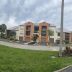 A two-story beige and orange apartment building with balconies, surrounded by green grass, palm trees, and cloudy skies. Construction cones and equipment are visible on the left and right sides of the image.
