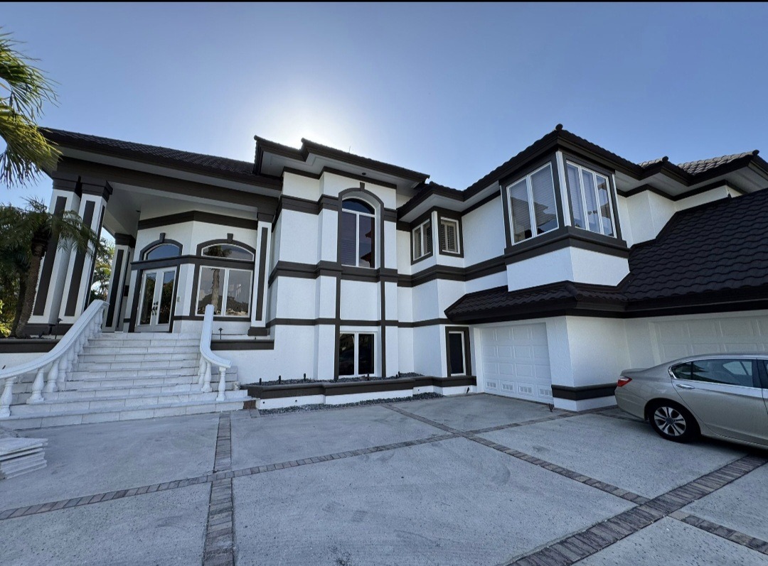 Objartel After Large, modern white house with dark trim, tall windows, a double staircase leading to the front entrance, a three-car garage, and a silver car parked in the driveway under a clear blue sky.