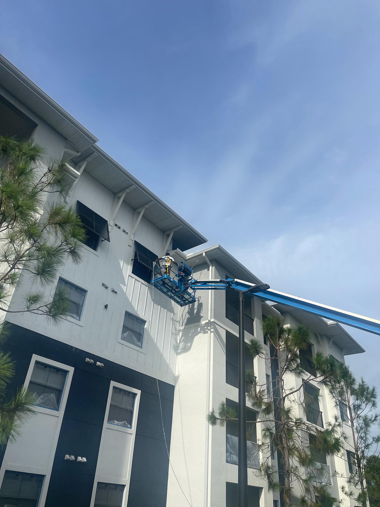 Two workers in a blue aerial lift are painting or repairing the upper exterior wall of a multi-story building on a clear day. The building has white walls with black accents and is surrounded by tall trees.