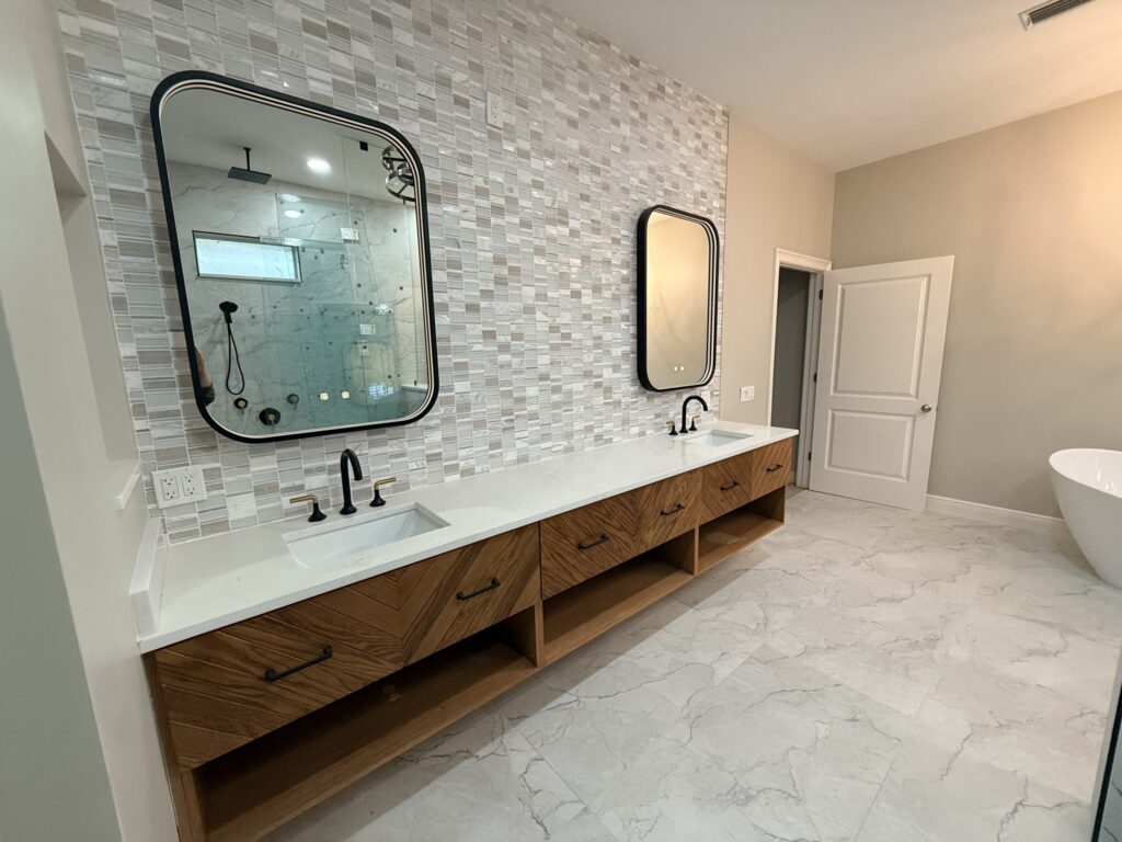 Modern bathroom designed by a remodeling contractor in Southwest Florida, featuring a double vanity with black fixtures, light wood drawers, mosaic tile backsplash, marble floor, and glass shower reflected in the mirror.