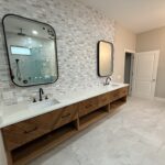 Modern bathroom designed by a remodeling contractor in Southwest Florida, featuring a double vanity with black fixtures, light wood drawers, mosaic tile backsplash, marble floor, and glass shower reflected in the mirror.
