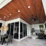 A modern covered patio with a wood-paneled ceiling, large black ceiling fan, dining table with wicker chairs, outdoor kitchen, and glass sliding doors, overlooking greenery outside.