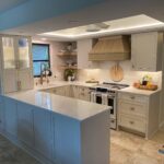 A modern kitchen with cream cabinets, a marble countertop island, a gas stove with a wooden range hood, under-cabinet lighting, and various kitchen essentials neatly arranged on the counters.