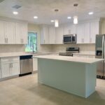 Modern kitchen with white cabinets, stainless steel appliances, an island with a light countertop, tile floor, pendant lights, and a window overlooking greenery.