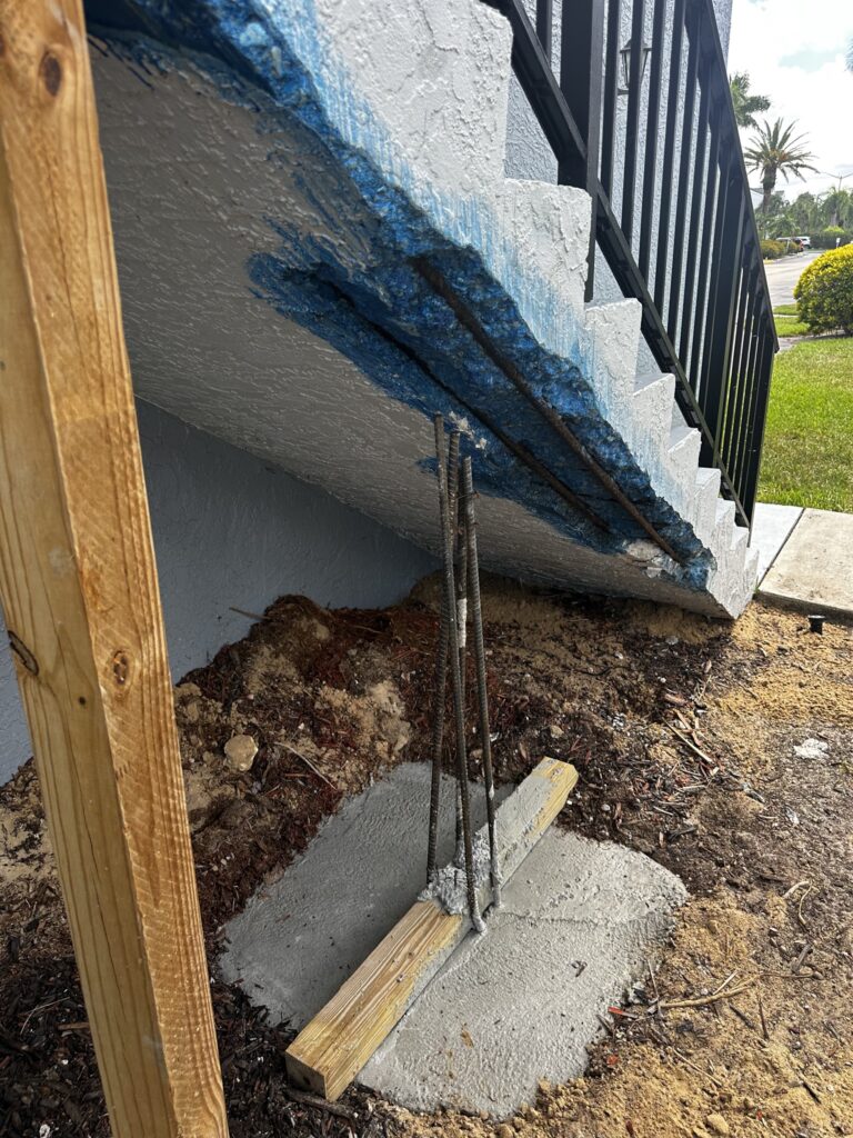 item_887ab371-08f8-46fd-918e-bdb135878061 A stairway with exposed blue-coated rebar underneath is supported by a wooden brace and a concrete base with vertical rebar rods, next to dirt and grass. A concrete restoration contractor in Florida could expertly handle such repairs.